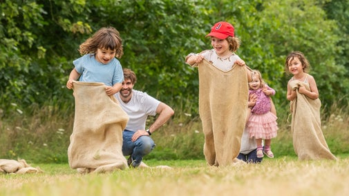 Three children in hessian sacks racing each other as two adults watch.